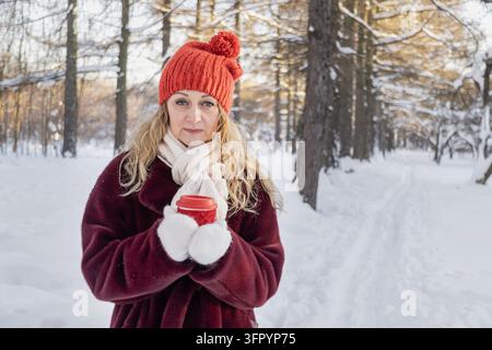 Mittlere ältere Frau hält einen wiederverwendbaren Kaffeebecher in einem verschneiten Wald Stockfoto
