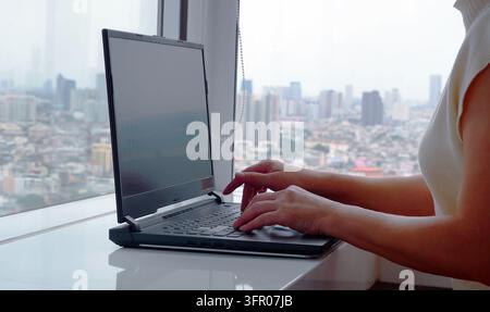 Nahaufnahme der Seitenansicht von weiblichen Händen, die auf einer weißen Tischplatte an einer Laptop-Tastatur arbeiten. Unerkennbare Frau, die am Computer tippt, vor einem unscharfen städtischen Hintergrund Stockfoto