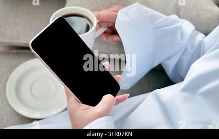 Weibliche Hand hält oder Smartphone mit einer Tasse schwarzen Kaffees auf Textil, digitalem NOMAD, freiberuflichem Arbeitsplatz oder Online-Shopping-Konzept verwendet Stockfoto