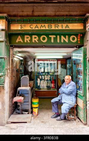 Ein alter Arrotino in einer blauen Arbeitsjacke sitzt auf einem Hocker an der offenen Tür einer traditionellen Messerschmiede in Siziliens Hauptstadt Palermo. Stockfoto