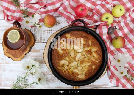 Oberansicht Flat Lay von hausgemachtem Apfelkuchen in einer Form, frischen Äpfeln, Tee mit Zitrone und weißen Blumen auf einem weißen Holztisch mit einem rot karierten Handtuch. Stockfoto