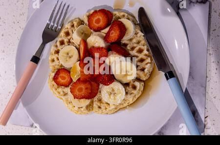 Ansicht von oben: Waffeln mit geschnittenen Erdbeeren, Banane und Sirup auf einem weißen Teller mit pastellfarbenen Besteckgriffen auf einer gesprenkelten Arbeitsplatte Stockfoto