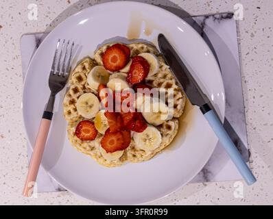 Ansicht von oben: Waffeln mit geschnittenen Erdbeeren, Banane und Sirup auf einem weißen Teller mit pastellfarbenen Besteckgriffen auf einer gesprenkelten Arbeitsplatte Stockfoto