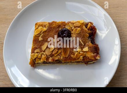 Rechteckiger Schnitt einer mit Mandeln gekrönten Bakewell-Tarte mit dunkler Kirschgarnitur und Fruchtfüllung, serviert auf einem weißen Keramikteller auf einem Holztisch Stockfoto