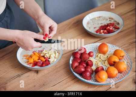 Frau schneidet Bananenscheiben in Müsli mit Erdbeeren und Aprikose, bereitet frisches Frühstück auf Holztablett zu, gesunde Zutaten und gesundes Ho Stockfoto