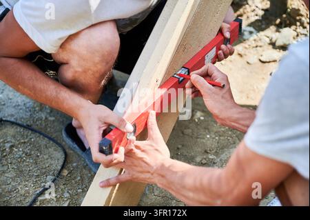 Arbeiter markieren einen Holzbalken mit Wasserwaage und Bleistift, um eine präzise Ausrichtung vor der Installation sicherzustellen. Bauarbeit im Freien, Teamarbeit, Genauigkeit und Konzept der Gebäudevorbereitung Stockfoto