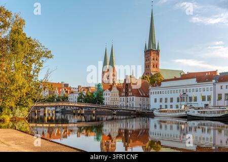 Uferpromenade in Lübeck im Norden Deutschlands mit historischen Hansestadtgebäuden, Kirchtürmen, einer Fußgängerbrücke und Spiegelungen auf dem Fluss. Stockfoto