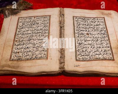 Seltene antike Quran-Handschrift mit handgeschriebener arabischer Kalligraphie auf offenen gealterten Pergamentseiten, die auf einem sattroten Tuch ruhen. Stockfoto