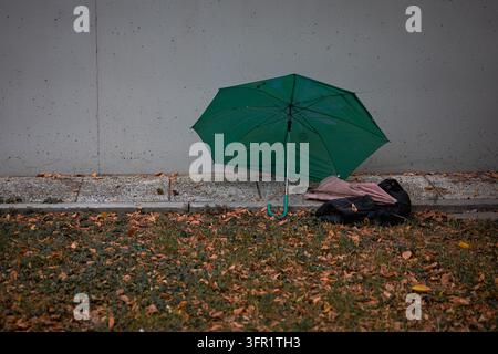 Verlassener grüner Regenschirm liegt auf dem Bürgersteig nahe einer Betonmauer an einem Herbsttag mit trockenen Blättern Stockfoto