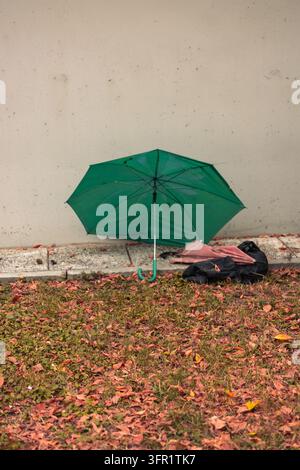 Verlassener grüner Regenschirm liegt auf dem Bürgersteig nahe einer Betonmauer an einem Herbsttag mit trockenen Blättern Stockfoto