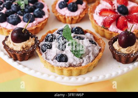 Verschiedene Mini-Fruchtkuchen mit Beeren und Sahne auf weißem Servierteller. Dessert-Hintergrund für süße Gebäckstücke, Partyessen und Feiern. Stockfoto