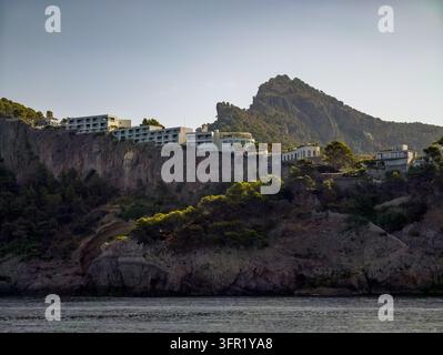 Horizontale Aufnahme von modernen Resort-Gebäuden, die am Rand schroffer Klippen thronen, umgeben von Kiefern, mit Blick auf das dunkelblaue Meer. Die Szene ist Stockfoto