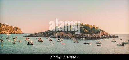 Zahlreiche Boote, darunter Segelboote und Motorboote, sind in einer ruhigen mediterranen Bucht in der goldenen Stunde in Mallorca, Spanien, vor Anker. Eine felsige, baumbewuchsene Stockfoto
