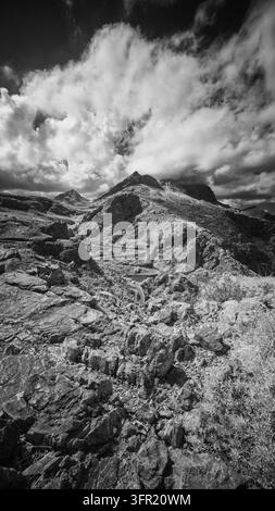 Eine vertikale Schwarz-Weiß-Fotografie zeigt eine gewundene Straße, die eine raue, felsige Berglandschaft in Mallorca, Spanien, durchquert. Dramatische Wolken füllen d Stockfoto