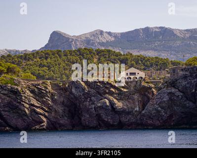 Häuser, die auf schroffen Felsen thronen, umgeben von üppigen grünen Kiefern, mit Blick auf das tiefe blaue Meer in Mallorca, Spanien. Im Hintergrund erheben sich majestätische Berge Stockfoto