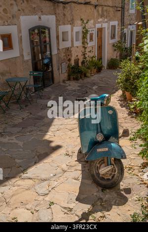 Ein klassischer, türkisfarbener Vespa-Roller steht auf einem sonnendurchfluteten, unebenen Steinpflaster in einer malerischen Dorfstraße in Mallorca, Spanien. Die antike Gebäudewand Stockfoto
