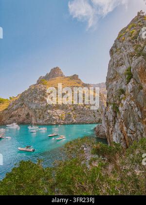Eine Luftaufnahme zeigt eine ruhige, türkisfarbene Wasserbucht, die sich zwischen schroffen, sonnenbeschienenen Felsklippen in Mallorca, Spanien, schmiegt. Mehrere Freizeitboote und Stockfoto