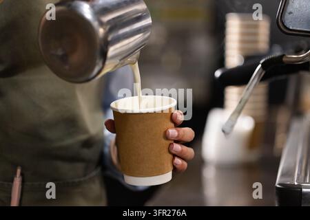 Asiatische Barista mit grüner Schürze gießt Milch in einen Pappbecher an der Kaffeetheke mit Stahlkanne. Espresso, Dampf, Café, Personal, schäumend, zum Mitnehmen, Tak Stockfoto