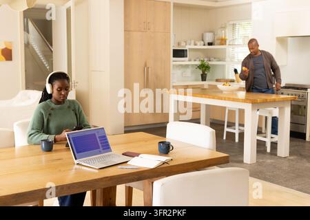 Paar in der Küche, Frau mit Kopfhörern und Pullover, die Laptop und Telefon benutzt, Mann hält Obstschale Stockfoto