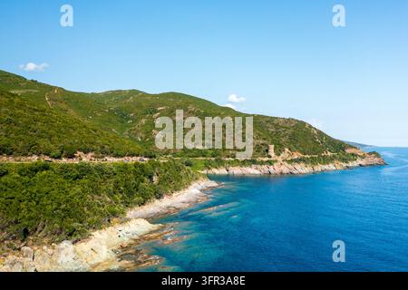 Felsige grüne Hügel treffen auf ein klares blaues Meer entlang der Küste bei Tour de l'Osse, mit einem kleinen historischen Turm in der Ferne. Stockfoto