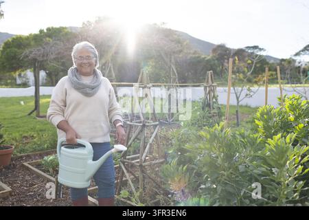 Reife afrikanische Frau steht im heimischen Garten im Gegenlicht und hält einen grünen Gießkannen mit Hochbeeten Stockfoto