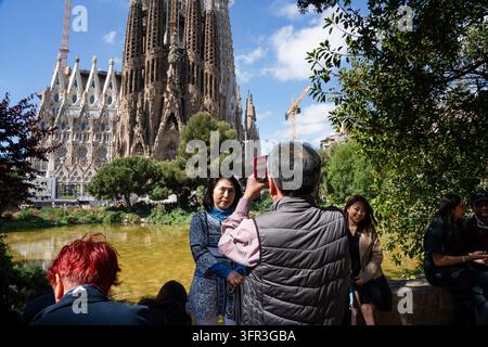 EIN FOTO VOR DER SAGRADA FAMILIA AUFNAHMEN, STRASSENFOTOGRAFIE-REPORTAGE, BARCELONA, SPANIEN, 2019: Ein Mann, von hinten gesehen, hält ein Mobiltelefon hoch, um ein Foto von einer Frau zu machen, die für ihn posiert, vor dem Hintergrund des Parksees und der Sagrada Familia. INFO: Barcelona ist eine lebendige spanische Küstenstadt, die für ihre ikonische Architektur, die mediterranen Strände, das historische Gotische Viertel, die reiche katalanische Kultur, Weltklasse-Fußball und eine geschäftige Großstadtatmosphäre voller Kunst, Essen und Musik bekannt ist. Stockfoto