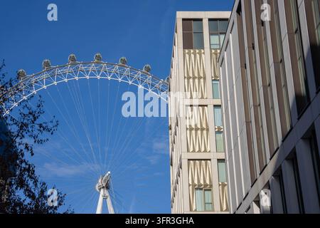 Blick auf das ikonische London Eye-Riesenrad, eingerahmt von modernen geometrischen Gebäuden am South Bank vor einem hellblauen Himmel. Stockfoto