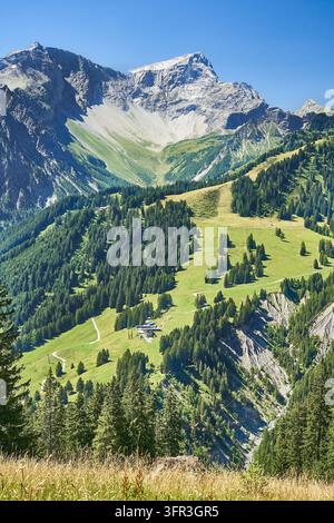 Eine Hochwinkelaufnahme zeigt eine kurvenreiche Straße, üppige grüne Hänge und dichte Wälder, die zu schroffen Berggipfeln unter einem klaren blauen Himmel führen. Stockfoto