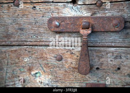 Rostige Eisenschnalle, die an einer verwitterten Holztür mit abblätternder Maserung, rauer Textur, alten Nägeln und gealterter Patina befestigt ist, Nahaufnahme von rustikalen Beschlägen und Stockfoto