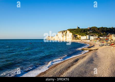 Eine weite Küstenansicht zeigt die Klippen, den Kiesstrand und die Küstenstadt Etretat im warmen Abendlicht, mit Booten, die am Ufer des Engländers angeschwemmt sind Stockfoto