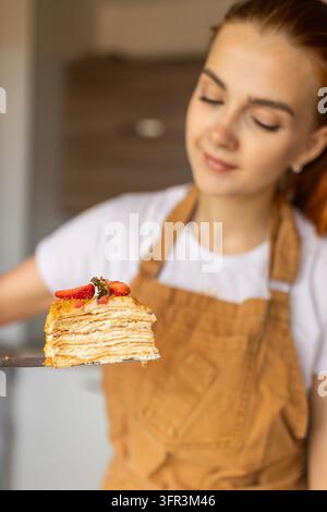 Glück auf der Scheibe. Frau in einer Schürze präsentiert eine Scheibe geschichteten Cremekuchens mit Erdbeeren in einer heimischen Küche. Stockfoto