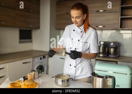 Ein verlockender Geschmack von Süße. Weibliche Patissière kostet Sahne von einem behandschuhten Finger, während sie während der Kuchenbereitung in der Küche einen Schneebesen hält. Stockfoto
