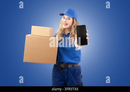 Fröhliche Lieferantin in blauer Uniform, die Pappkartons hält und der Kamera einen leeren Smartphone-Bildschirm auf blauem Hintergrund zeigt. Perfec Stockfoto