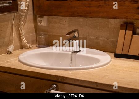 Weißes Keramik-Waschbecken mit einem Chrom-Wasserhahn, eingebaut in eine beige Stein-Arbeitsplatte, mit einem wandmontierten Telefon und einem Trinkglas im Hintergrund Stockfoto