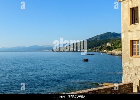 Ruhiges Meer mit der Dorfküste, felsiger Küste und Bergen im Hintergrund. Ein sonnenbeschienenes Steingebäude rahmt die rechte Seite des Bildes ein. Stockfoto