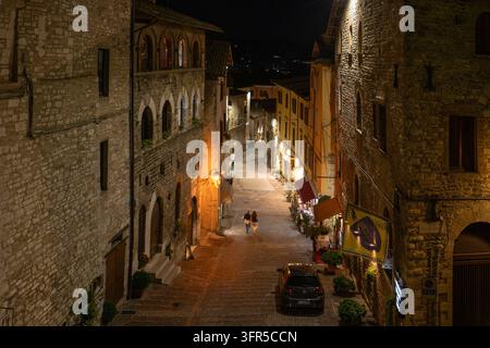 Spaziergänger und ein Auto in einer engen, beleuchteten Altstadtgasse zwischen hohen Steinhäusern, Gubbio, Umbrien, Italien Stockfoto