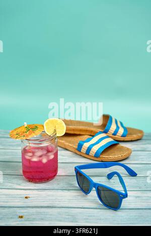 Erfrischendes rosa farbenes Getränk mit einem Schirmchen und einer Zitronenscheibe, blauer Sonnenbrille und Strandsandalen, auf einem Holztisch und grünem Hintergrund Stockfoto
