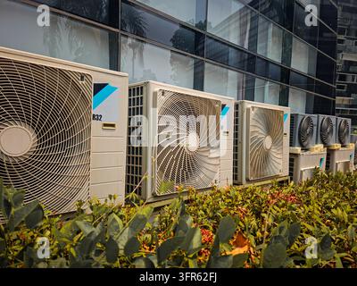 Mehrere mobile Klimaanlagen stehen neben einem modernen Bürogebäude aus Glas, mit gepflegten Pflanzen im Vordergrund. Die Szene veranschaulicht c Stockfoto