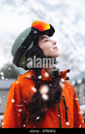 Lächelnde junge Frau, Porträt, stehend und genießt den Schneefall, trägt Schutzkleidung, Helm und Schutzbrille. Wintersportort Stockfoto