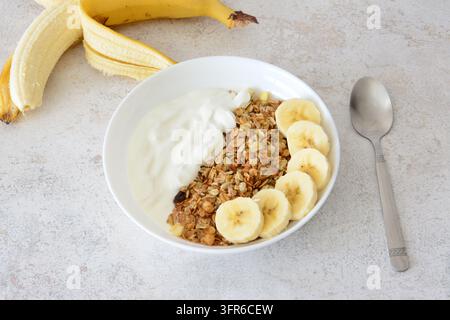 Eine Frühstücksschüssel mit Granola, Joghurt und Bananenscheiben sowie Bananenfrucht mit Schale Stockfoto