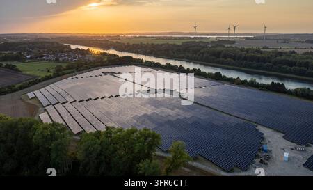 Luftaufnahme einer großen Solarfarm mit Windturbinen im Hintergrund, vor einer malerischen Landschaft an einem Kanal bei Sonnenuntergang. Solarpanele umgeben Stockfoto