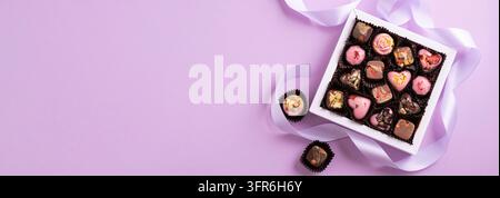 Eine Auswahl an handgemachten Schokoladen und Trüffeln in einer weißen Geschenkschachtel mit einer violetten Schleife auf einem pastellfarbenen Hintergrund ist ein süßes Geschenk Stockfoto