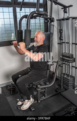 Seitenansicht eines konzentrierten älteren Mannes mit grauem Bart, der auf einem Heim- oder Studio-Multifunktions-Fitnessgerät trainiert. Er führt einen Brustdruck oder Stockfoto