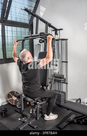 Rückansicht eines aktiven Senior Mannes mit grauen Haaren, der auf einem Multifunktions-Heimfitnessgerät trainiert. Er führt eine Latzugübung aus, um zu stärken Stockfoto