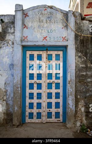 Verwitterte blau-weiße Holztür mit Metallnieten unter einer Wand mit einer alten gemalten Inschrift, Gujarat, Mandvi, Indien Stockfoto