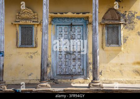 Eine geschnitzte hellblaue Holztür steht zwischen zwei verzierten Gitterfenstern an einer gelben Fassade, Gujarat, Mandvi, Indien Stockfoto