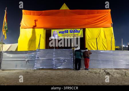 Zwei Männer stehen an einem Metallzaun vor einem großen orangefarbenen und gelben Zelt mit einem Salbeiporträtbanner während der Maha Kumbh Mela in Uttar Pradesh, Prayagraj, Indien Stockfoto