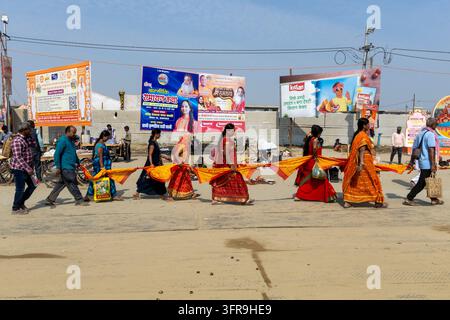 Eine Reihe von Frauen in farbenfrohen Saris und Männer tragen ein langes orangefarbenes und gelbes Tuch entlang einer sandigen Straße an Plakaten vorbei, Uttar Pradesh, Prayagraj, Indien Stockfoto