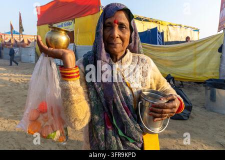 Lächelnde ältere Frau hält einen Messingtopf und einen Beutel mit Opfergaben in einem Zeltlager auf der Maha Kumbh Mela, Uttar Pradesh, Prayagraj, Indien Stockfoto
