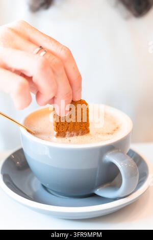 Hand einer Frau taucht einen karamellisierten Keks in einen schaumigen Chai Latte, der in einer grauen Tasse mit Untertasse serviert wird, daneben liegt ein goldener Löffel. Nahaufnahme-Lebensstil s Stockfoto